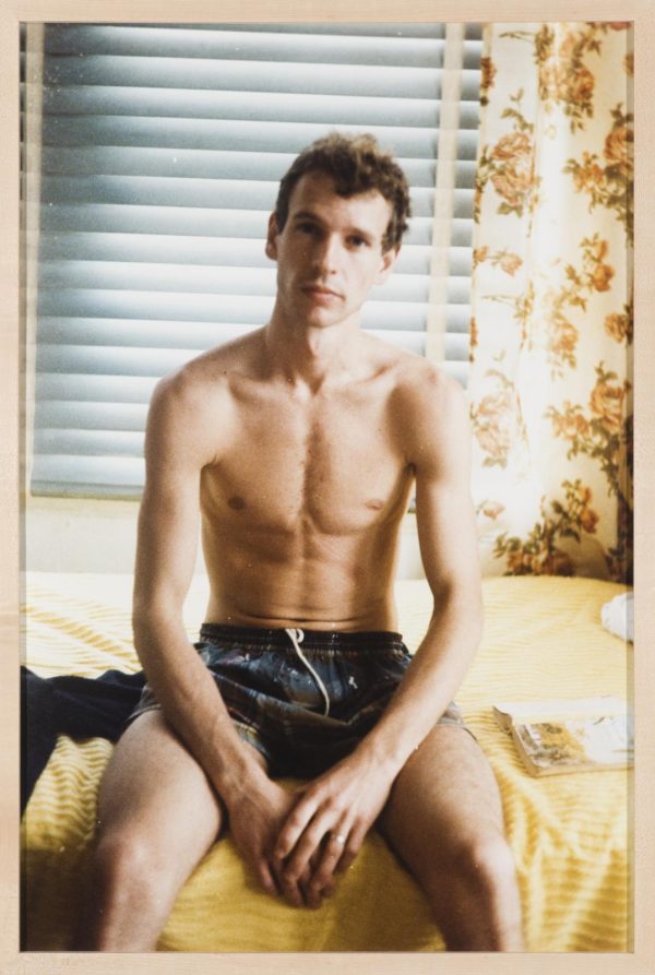 A topless, light-skinned man sits on a bed in a swimming suit. His head tilts just slightly to his left as he looks directly at the viewer.
