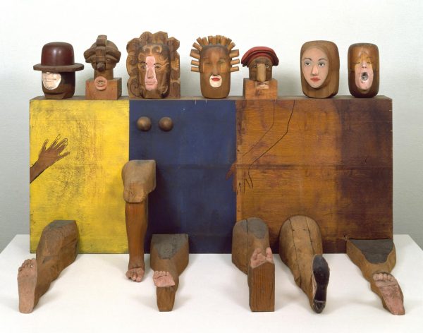 A block sculpture painted yellow blue and natural brown with seven heads and six legs attached to it, and one painted hand on it.
