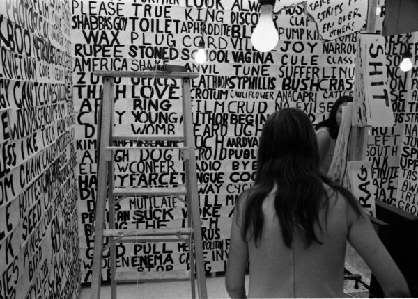 Black and white photo of a person with long hair in a room with writing writing covering the walls from the floor to the ceiling. A ladder stands to their left and bare bulbs hang from the ceiling.