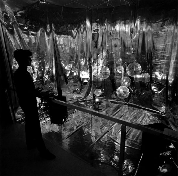 Black and white image of a person looking at a room from behind a stantion filled with clear orbs hanging from the ceiling with shimmery fabric on the walls and floor