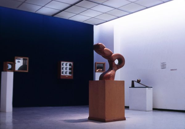An abstract wooden sculpture resembling hands making a bunny shadow on a pedestal in a room full of other artworks. One wall is painted royal blue.