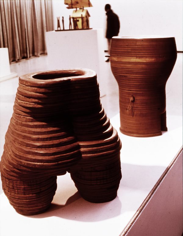A terracotta sculpture of a lower torso and butt seemingly formed from coils on a white pedestal with other sculptures