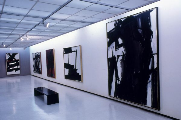 Five large paintings, predominately black and white, hang along a gallery wall.