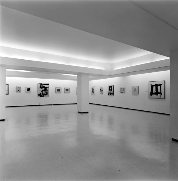 Many small black and white paintings hang along a gallery wall.