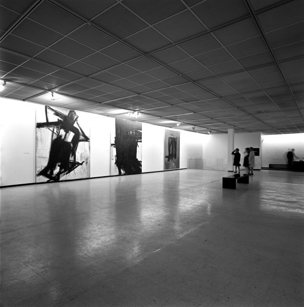 Visitors admire three large black and white paintings hanging against a gallery wall.