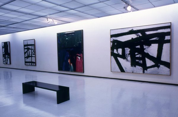 Three black and white and one multicolored paintings hang along a gallery wall.