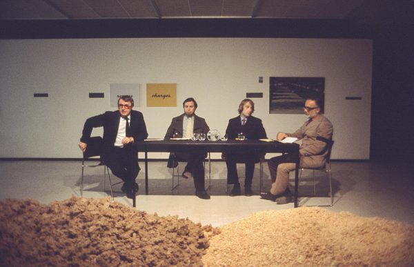 Four men sit at a table in a gallery space. In front of them are two piles of grain or sand