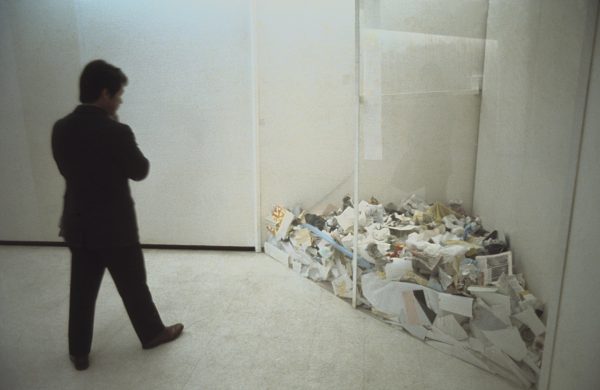 A person looks down at a pile of loose paper that has been relegated to the corner of a room, trapped by a clear divider
