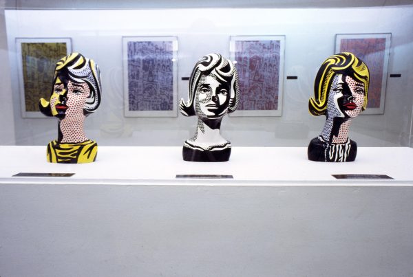 Three female busts painted in a pop style rest on a long white pedestal in a gallery with several paintings hung on the wall behind it