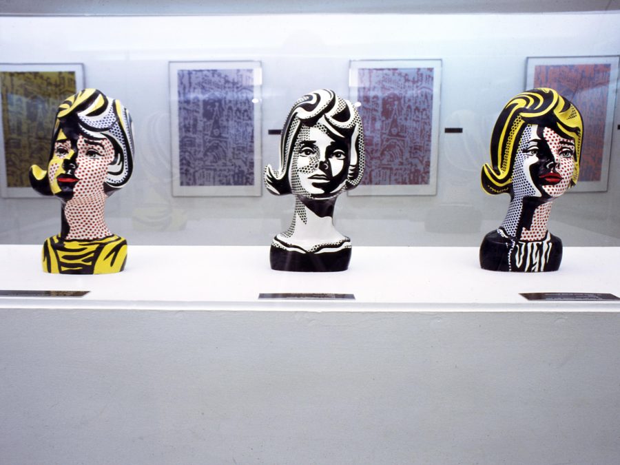 Three female busts painted in a pop style rest on a long white pedestal in a gallery with several paintings hung on the wall behind it