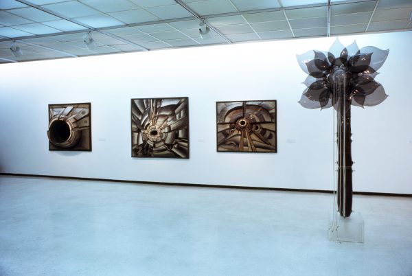 Installation of three 3D rectangular formations hanging on a white wall next to a tall sculpture of a flower with the shadow of its petals as it's captured mid-movement