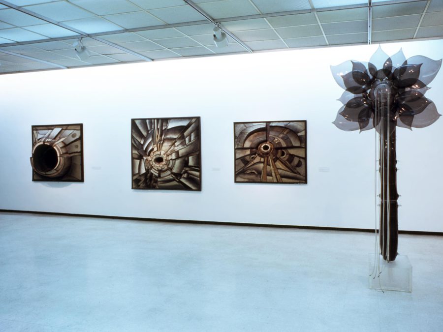Installation of three 3D rectangular formations hanging on a white wall next to a tall sculpture of a flower with the shadow of its petals as it's captured mid-movement