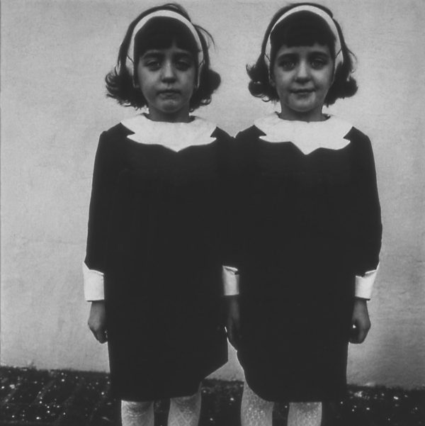 A black-and-white portrait of twins standing shoulder to shoulder. The twins are adorned in identical black dresses with white collars, white headbands, and white stockings.