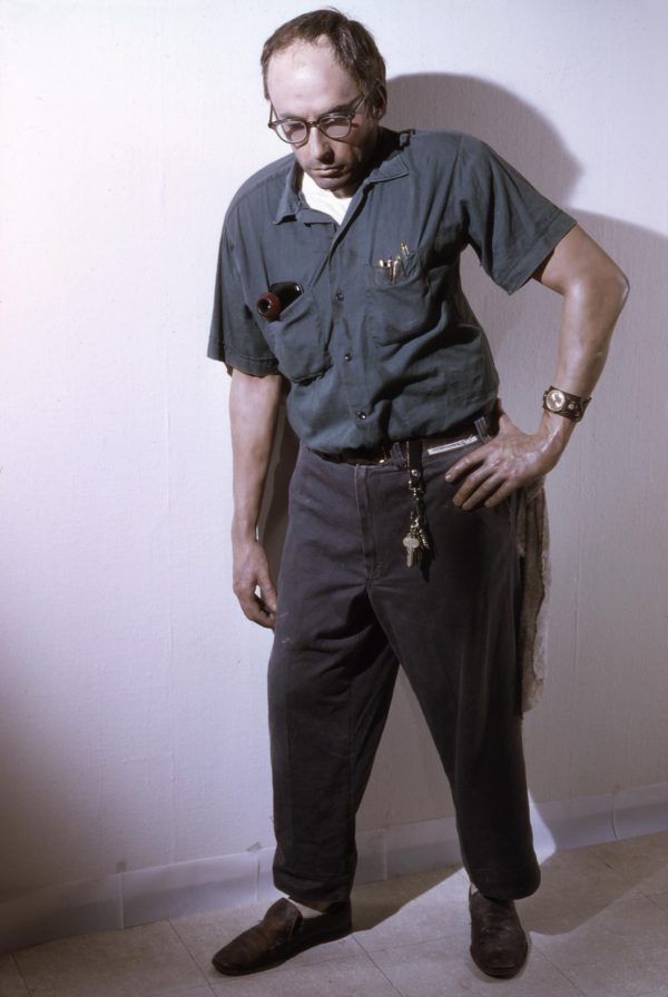 Life-size sculpture of a man dressed as a janitor, who leaning against the wall with his left hand on his hip. He has tools and pens in his shirt pockets, and keys attached to his belt loop.