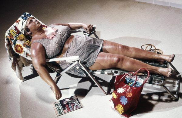 A female-presenting person in a swimsuit lounging with their eyes closed on a folding chaise lounge with a red beach tote with flowers in it and a magazine on the ground