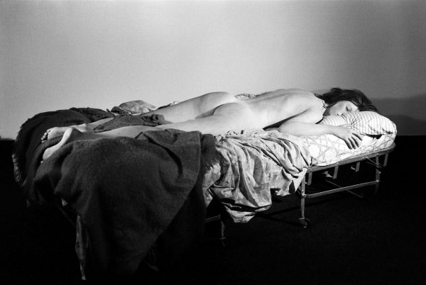 A nude person lying on a pull-out bed, the covers messy beneath them