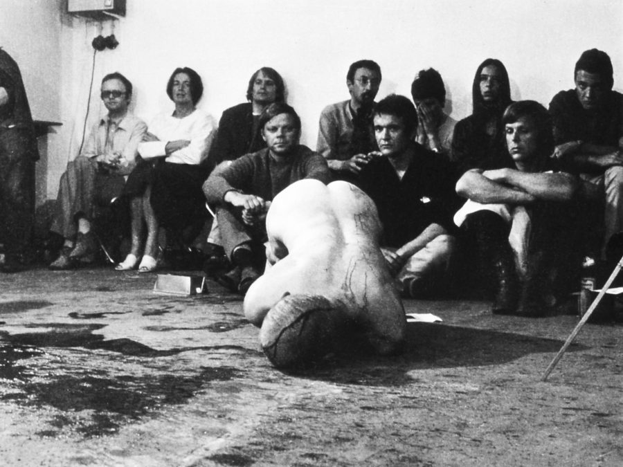A naked person in a child's post with a dark liquid dripping from their head in front of a group of people sitting in observation