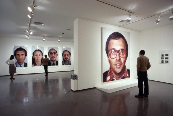 A gallery installation shows people viewing large-scale portraits of faces, four in the rear and one in the foreground.