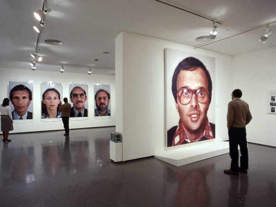 A gallery installation shows people viewing large-scale portraits of faces, four in the rear and one in the foreground.