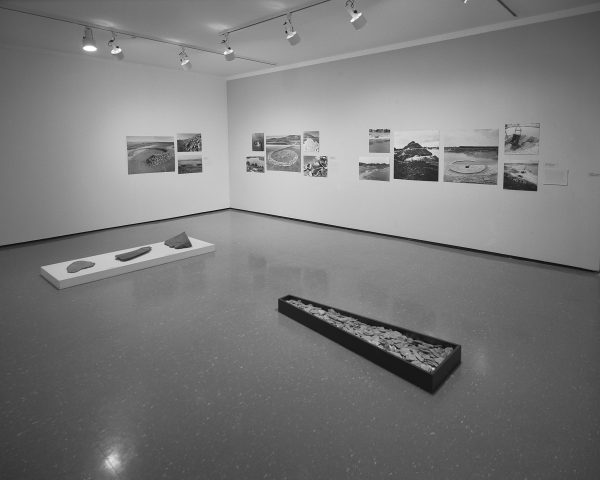 Black-and-white installation view of photographs and two sculptures that feature rocks