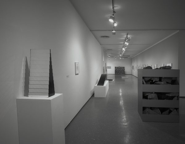 Black-and-white installation view of sculptures, including a stepped pyramid form and a crate-like sculpture filled with rocks