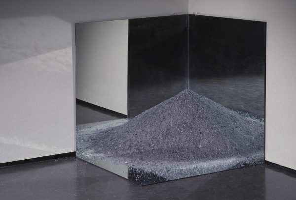 A sculpture composed of mirrors and a mound of sand appears in the corner of an art installation.