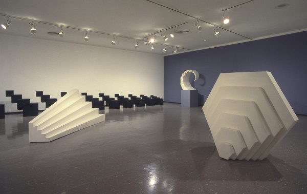 Four sculptures of various repeating shapes, three white, one black, in a gallery space with one wall painted purple