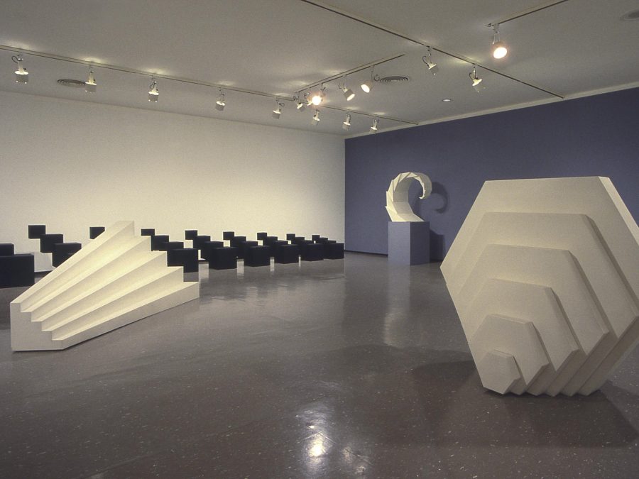 Four sculptures of various repeating shapes, three white, one black, in a gallery space with one wall painted purple