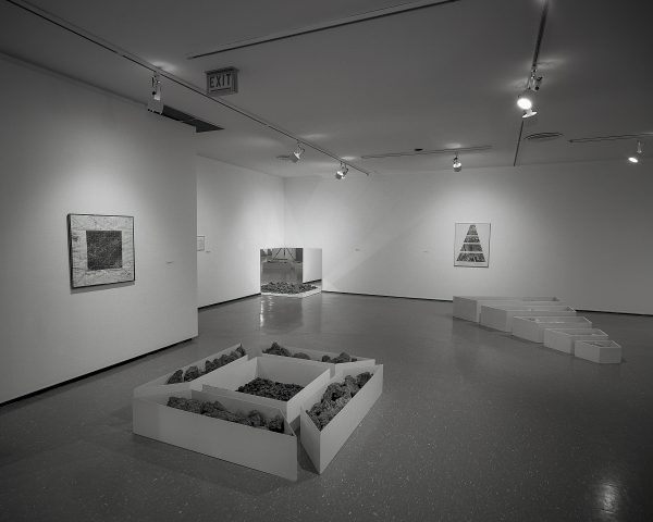 A black-and-white installation view shows photographs and sculptures made of geometric containers filled with stones.