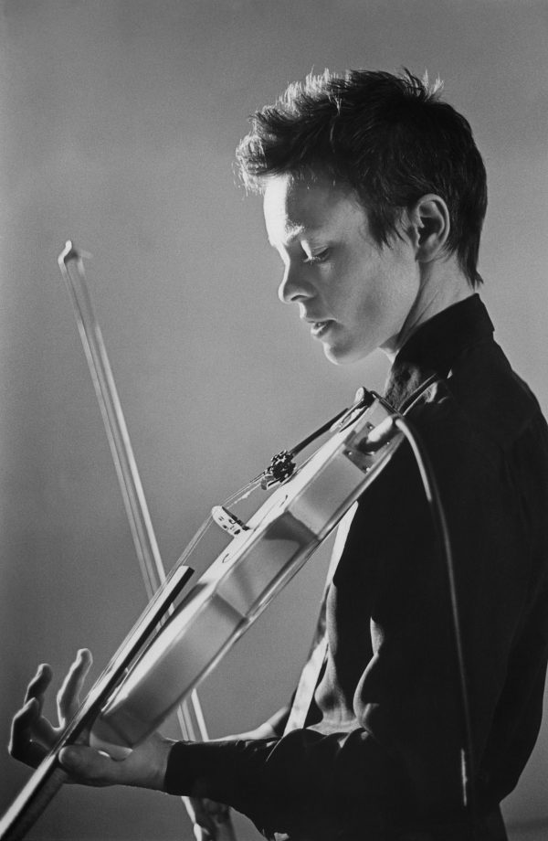 A black-and-white photograph of a youthful person with eyes closed holds a violin and bow, ready to play