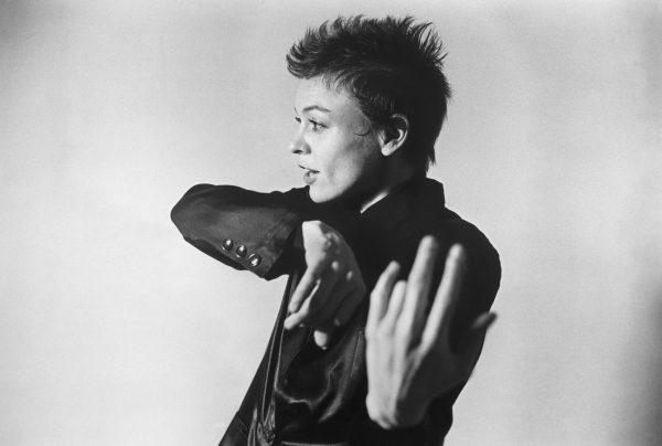 A short-haired person is shown in a black and white portrait of her profile, her arms gracefully held to the side and her mouth slightly opened, as though in the midst of saying something.