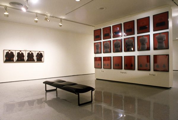 A gallery space with an installation of a series of images displayed in a three by six grid on the right wall and four images hung side by side in a row on the left.