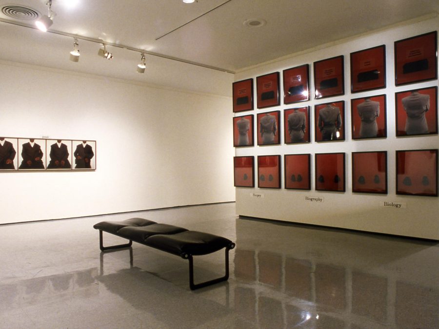 A gallery space with an installation of a series of images displayed in a three by six grid on the right wall and four images hung side by side in a row on the left.