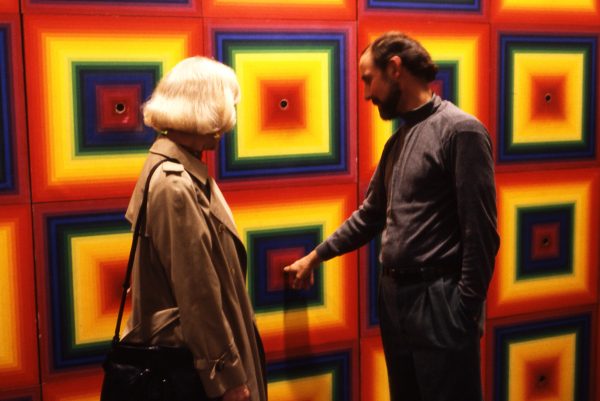 A man and a woman stand in front of a wall with a colorful, square, radial gradients with holes at the center of each. The man has finger in one hole.