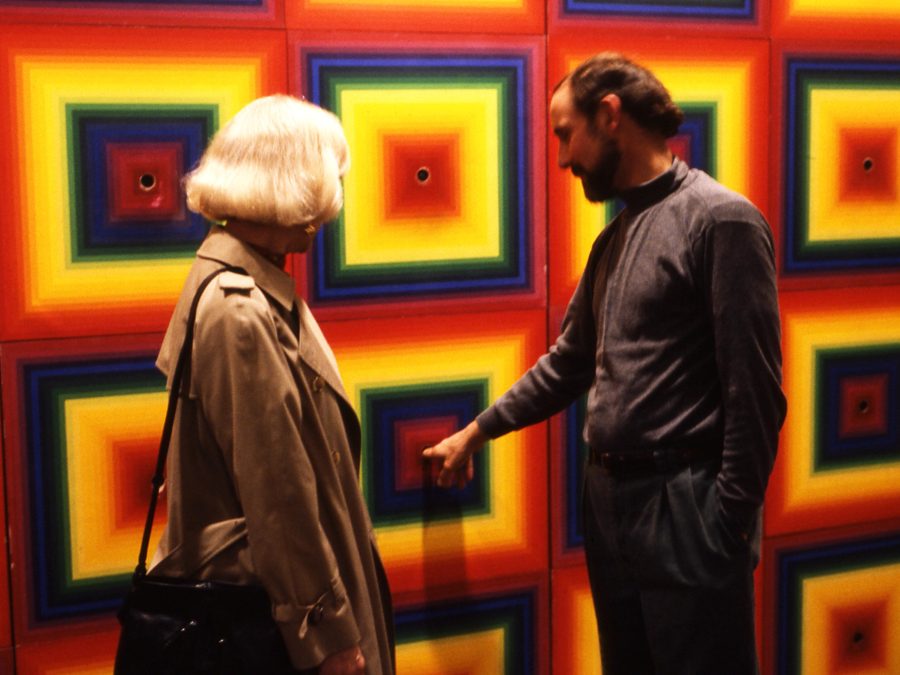 A man and a woman stand in front of a wall with a colorful, square, radial gradients with holes at the center of each. The man has finger in one hole.