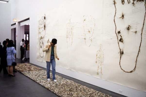 A man walks tentatively across a rectangular stone path installed in a gallery. On the wall beside him are images of feet which have been punctured.