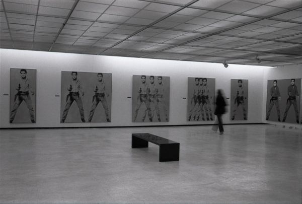 A black-and-white photograph of a gallery with several large paintings of Elvis Presley with dark hair in Western clothing holding a handgun. A black bench sits in the center of the room. A blurry person moves in the gallery.