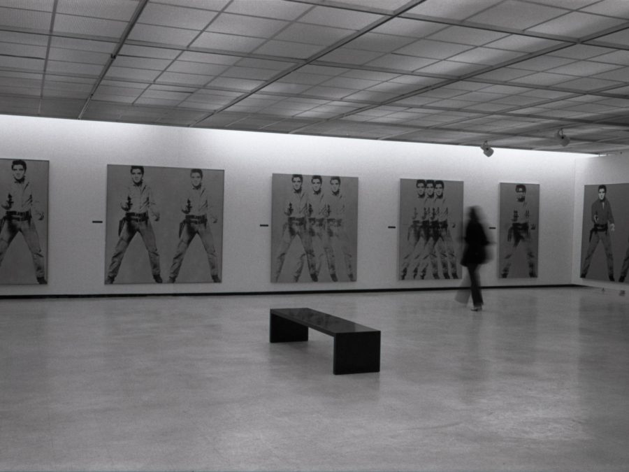 A black-and-white photograph of a gallery with several large paintings of Elvis Presley with dark hair in Western clothing holding a handgun. A black bench sits in the center of the room. A blurry person moves in the gallery.
