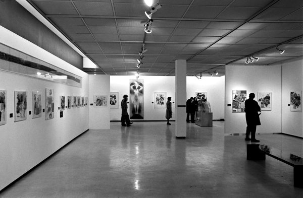A black-and-white installation photograph of graphic artworks and visitors.