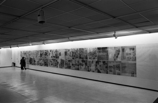 A black-and-white photo shows an installation view of dozens of graphic artworks. Two people view the works.