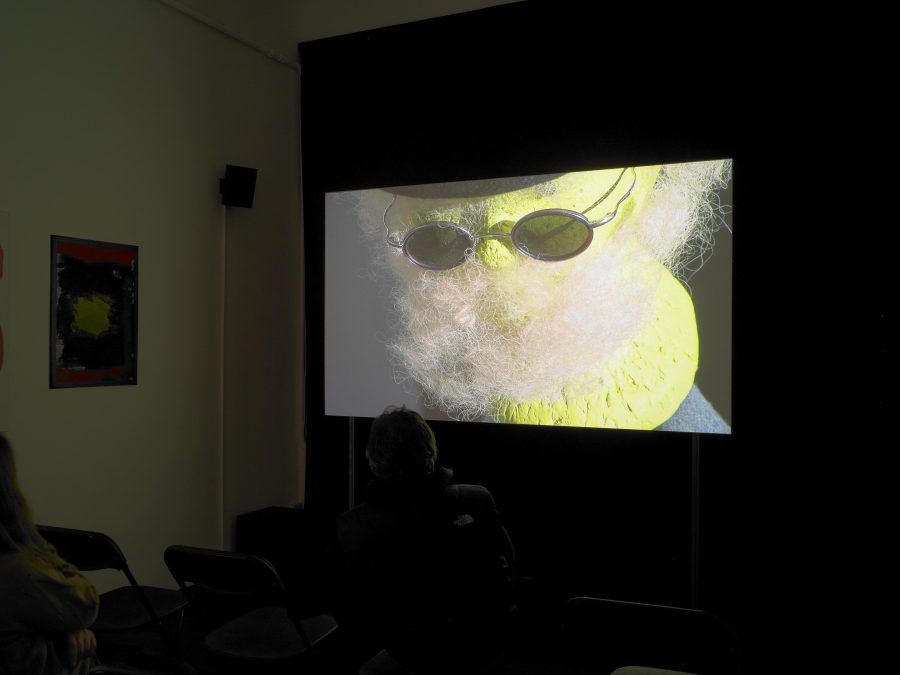 Installation view of video projection: a bright yellow object with sunglasses and tan fuzz that resembles a beard