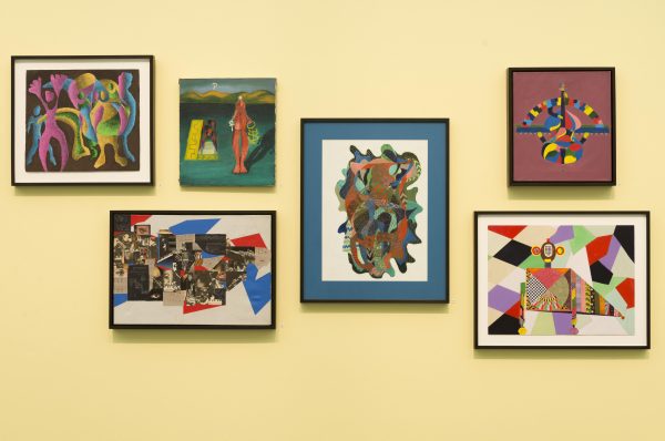 Six colorful paintings featuring geometric forms in bright colors are hung in a cluster on a yellow wall.