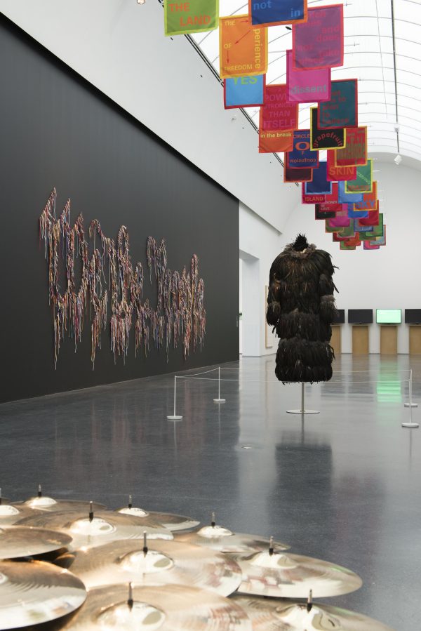 A feathered coat is displayed in the middle of a gallery, cymbals are in the foreground, and 
