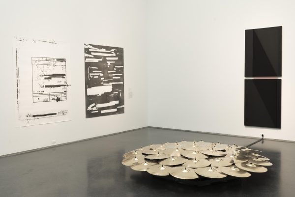 Several dozen brass cymbals are clustered together in a circle. Behind them to the left, two black-and-white paintings hang side by side, and to the right, two black panels are stacked vertically.