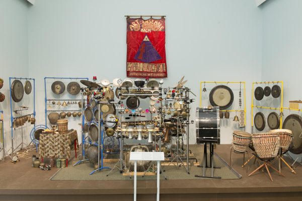 An eclectic array of percussion instruments fills a stage. Above them hangs a red banner featuring a pyramid and the words