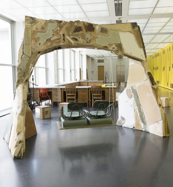 A two-seat swing made of school chairs hangs from an arch constructed from found materials crudely patched together.