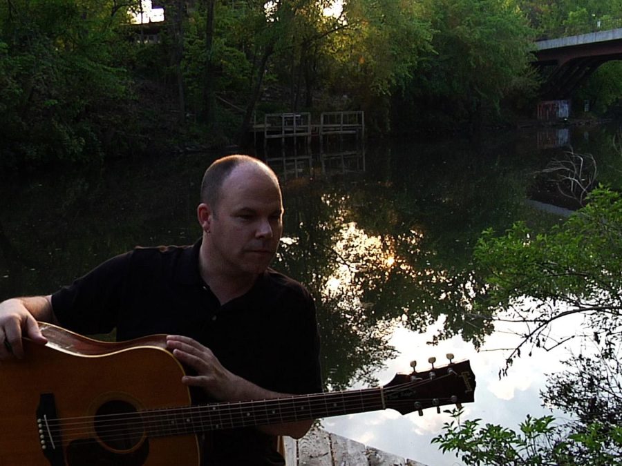 A man holding a guitar in front of a secluded tree-lined body of water