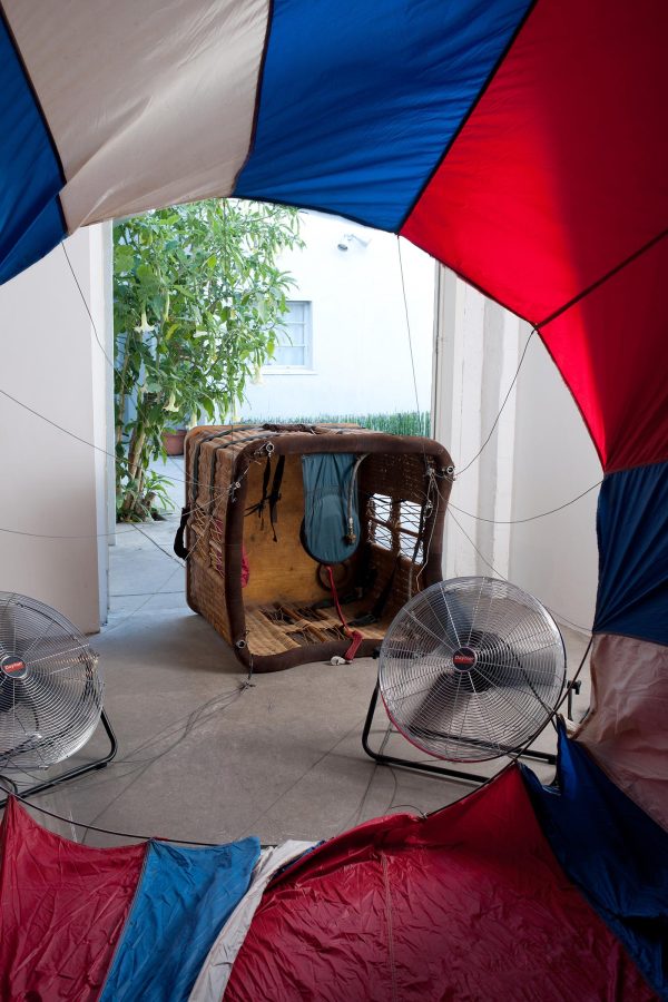 View through a circular opening in red, white, and blue fabric toward two fans and a large basket with strings attached to the fabric