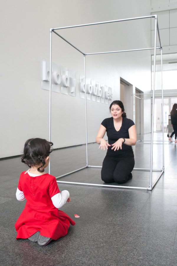 A woman kneels in an open-sided cage facing a girl who mimics her posture