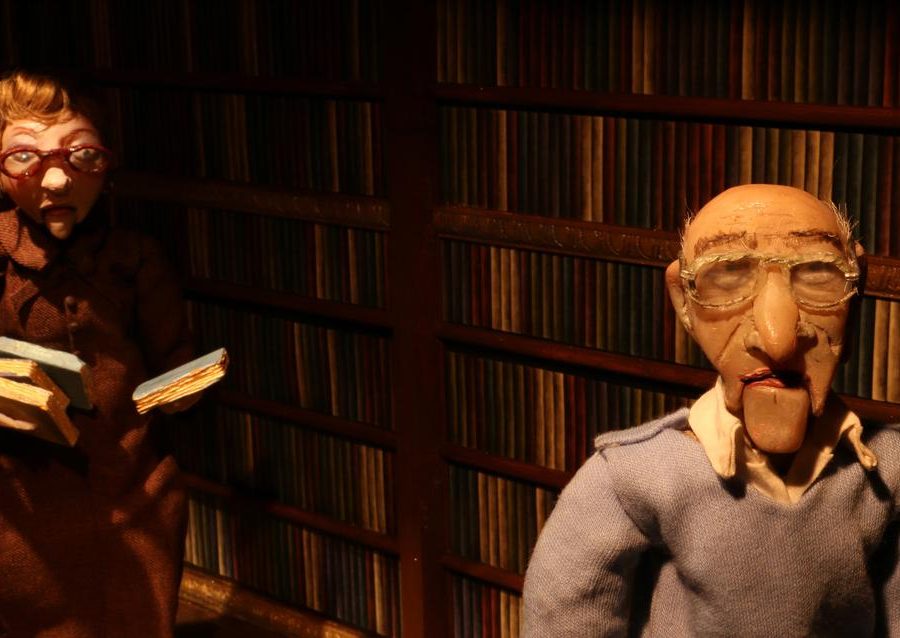 Puppets of an old man and woman walk through a dark room past a wall lined with books.